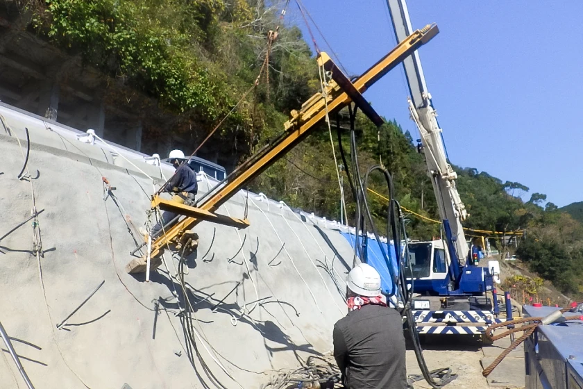 クレーンで吊るした削孔機を使用して斜面に穴を開ける作業員