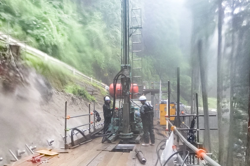 雨天の中、山間部で削孔作業を行う作業員たちの様子